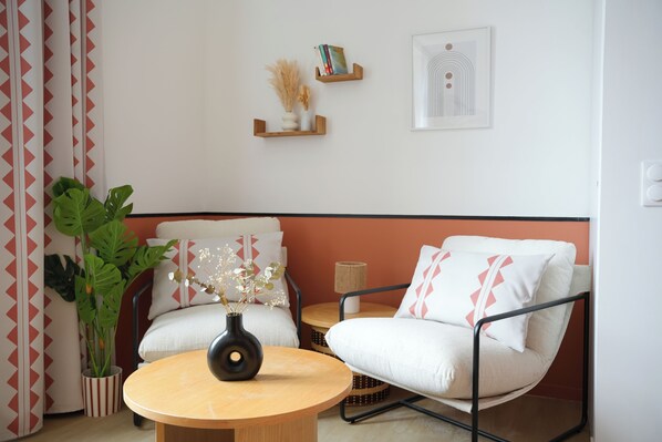Una habitación con una mesa redonda de madera, dos sillones blancos, una planta verde grande y un estante en la pared con libros y objetos decorativos.