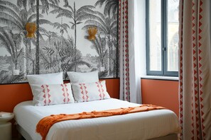 A bed with white bedding and orange accents, a wall with a tropical plant pattern, and a window with curtains.