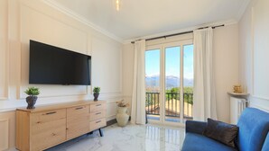 A modern living room with a flat-screen TV, a blue sofa, a wooden cabinet, and a large window with a balcony.