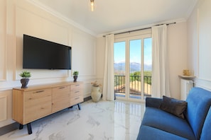 A modern living room with a flat-screen TV, a blue sofa, a wooden cabinet, and a large window with a balcony. - Black Pepper Stresa (Stresa)