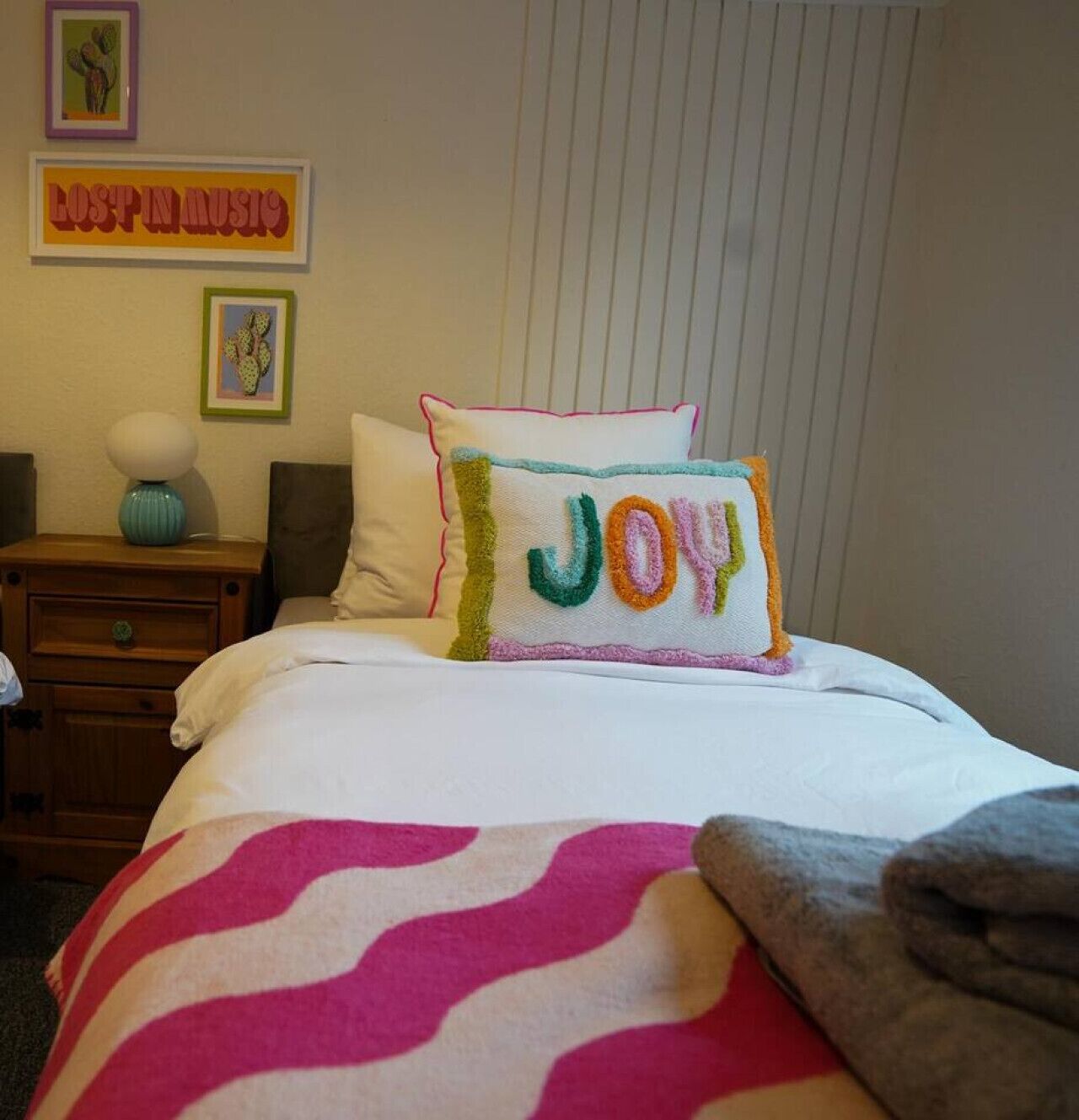 A neatly made bed with a colorful pillow that reads "JOY" and a decorative pillow with a cactus design. There is a small table with a lamp and a framed picture on the wall.