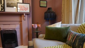 A cozy living room with a fireplace, a green sofa with patterned cushions, a small table with a vase of flowers, and a wall-mounted lamp. - The Maple House common views, central Lincoln (Lincolnshire)