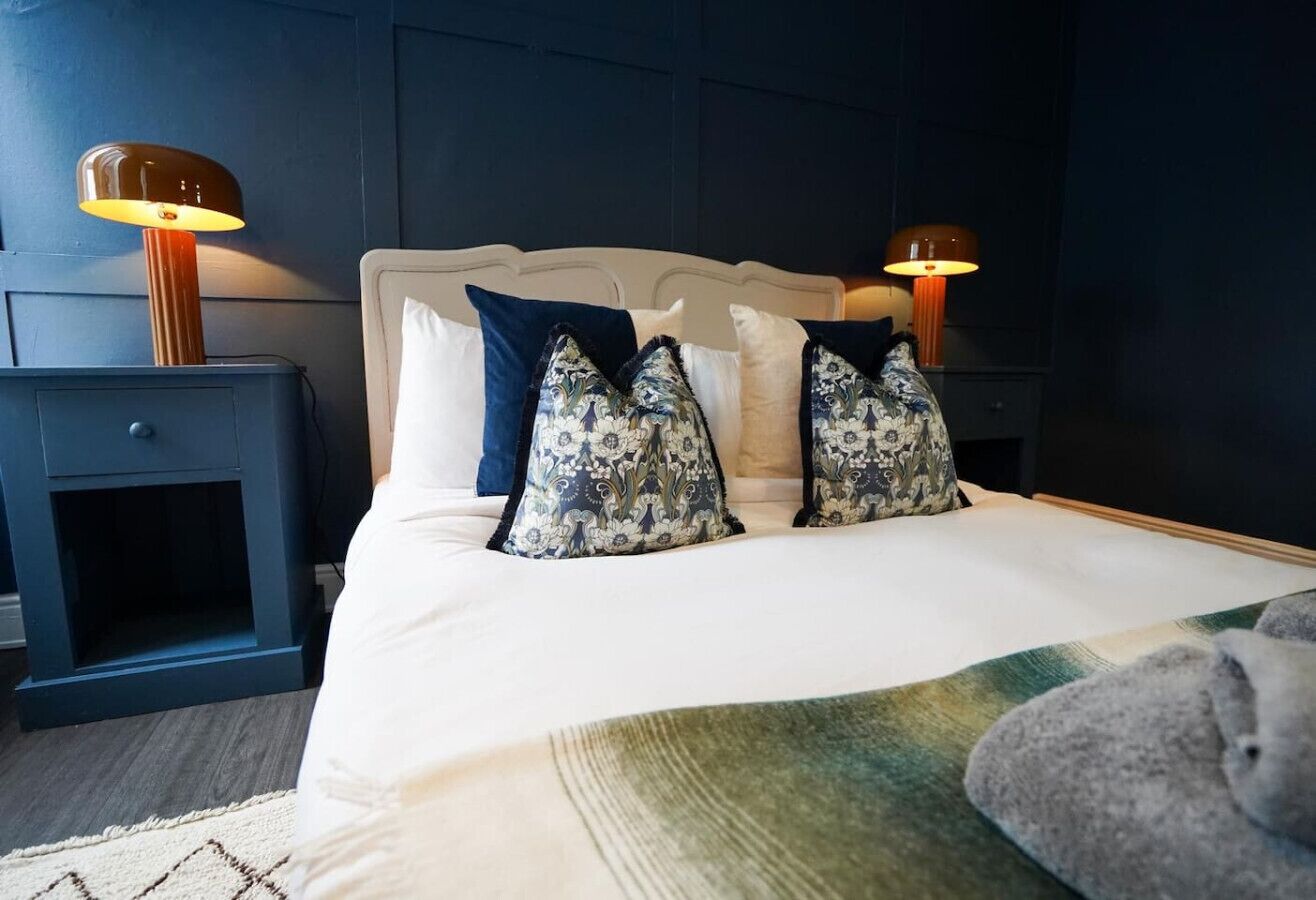 Un lit bien fait, agrémenté d’oreillers à motifs, une table de chevet avec une lampe et un mur de couleur sombre.