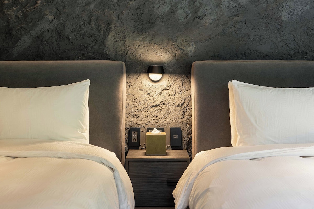 Een slaapkamer met een structuurmuur van steen, twee bedden met witte lakens, een nachtkastje met een lamp en een klein tafeltje met een geel voorwerp.