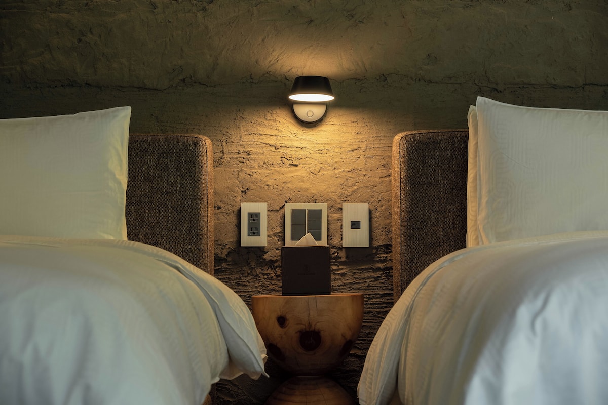 Een kamer met twee bedden, een houten kruk, een wandlamp en een verdeeldoos.