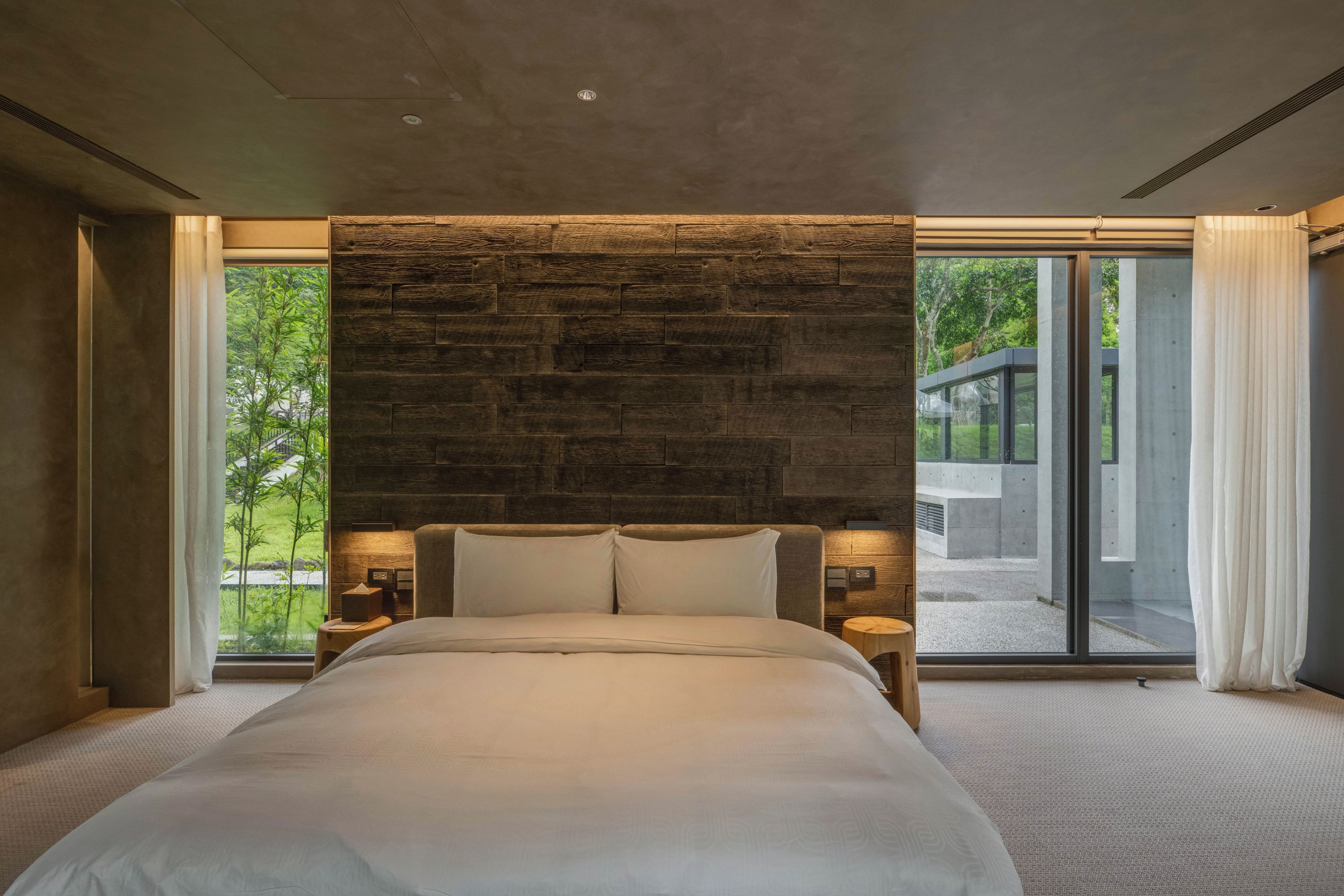 A modern bedroom with a large bed, stone wall, and large windows.