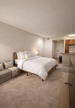 A bedroom with a bed, a nightstand, a lamp, a sofa, and a kitchen area with cabinets and a refrigerator. - Cozy 1-bedroom Marina Living (Marina del Rey)