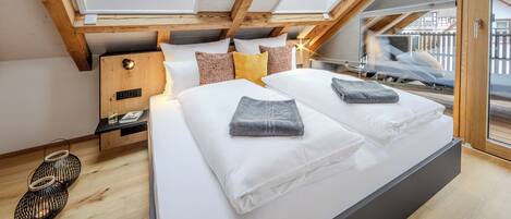 Un dormitorio moderno con tragaluz, vigas de madera, una cama grande con ropa blanca y una mesita de noche con lámpara.