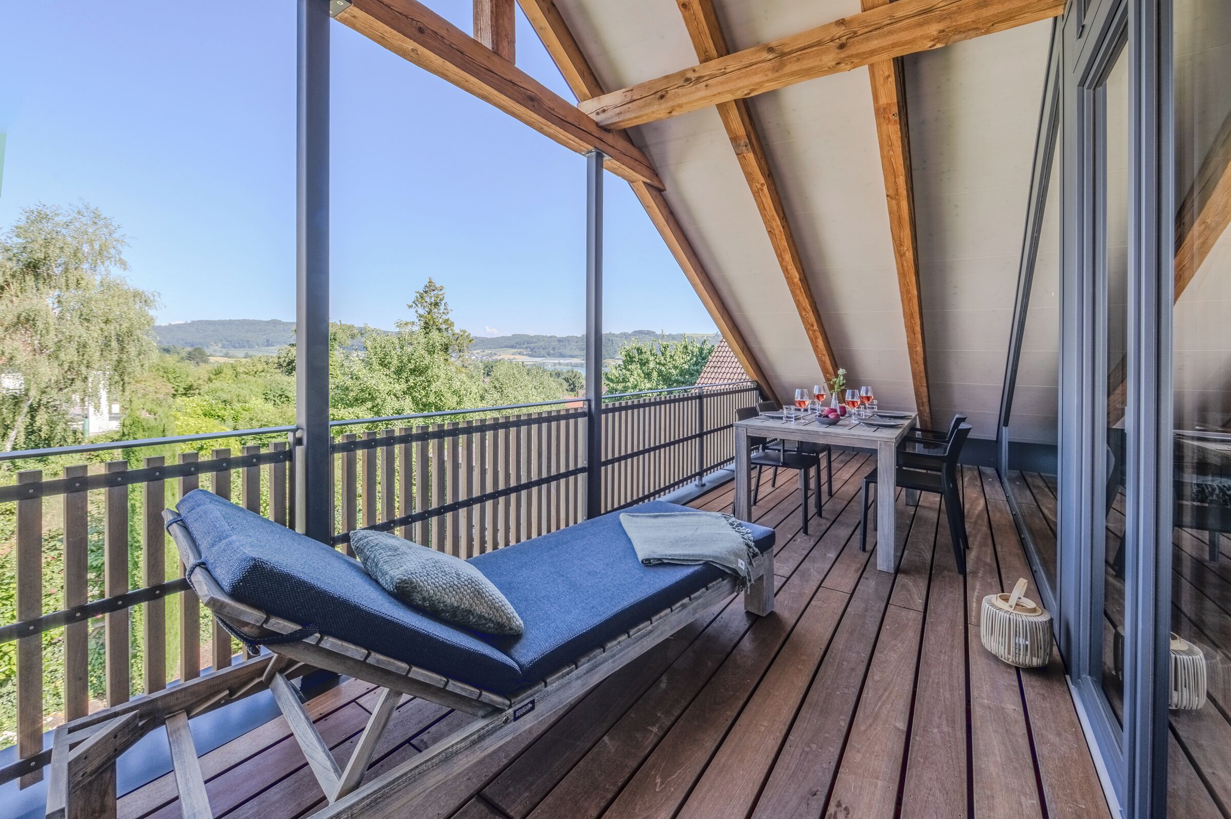 Ein Balkon mit Holzboden, einem Tisch mit Stühlen und einem Liegestuhl mit Kissen.