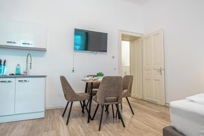 A compact kitchen with white cabinets, a dining table with four chairs, a flat-screen TV mounted above the table, and a door leading to another room. - Comfortable 1BR Apartment Long Stays (Vienna)