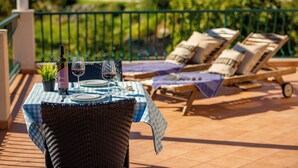 Een eettafel buiten, met rieten stoelen, een groen-wit geruit tafelkleed en uitzicht op een tuin met een groene hekken.'