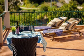 A dining table set outdoors with wicker chairs, a green and white checkered tablecloth, and a view of a garden with a green fence. - MOLUNAT DUBROVNIK (Gornji Brgat)