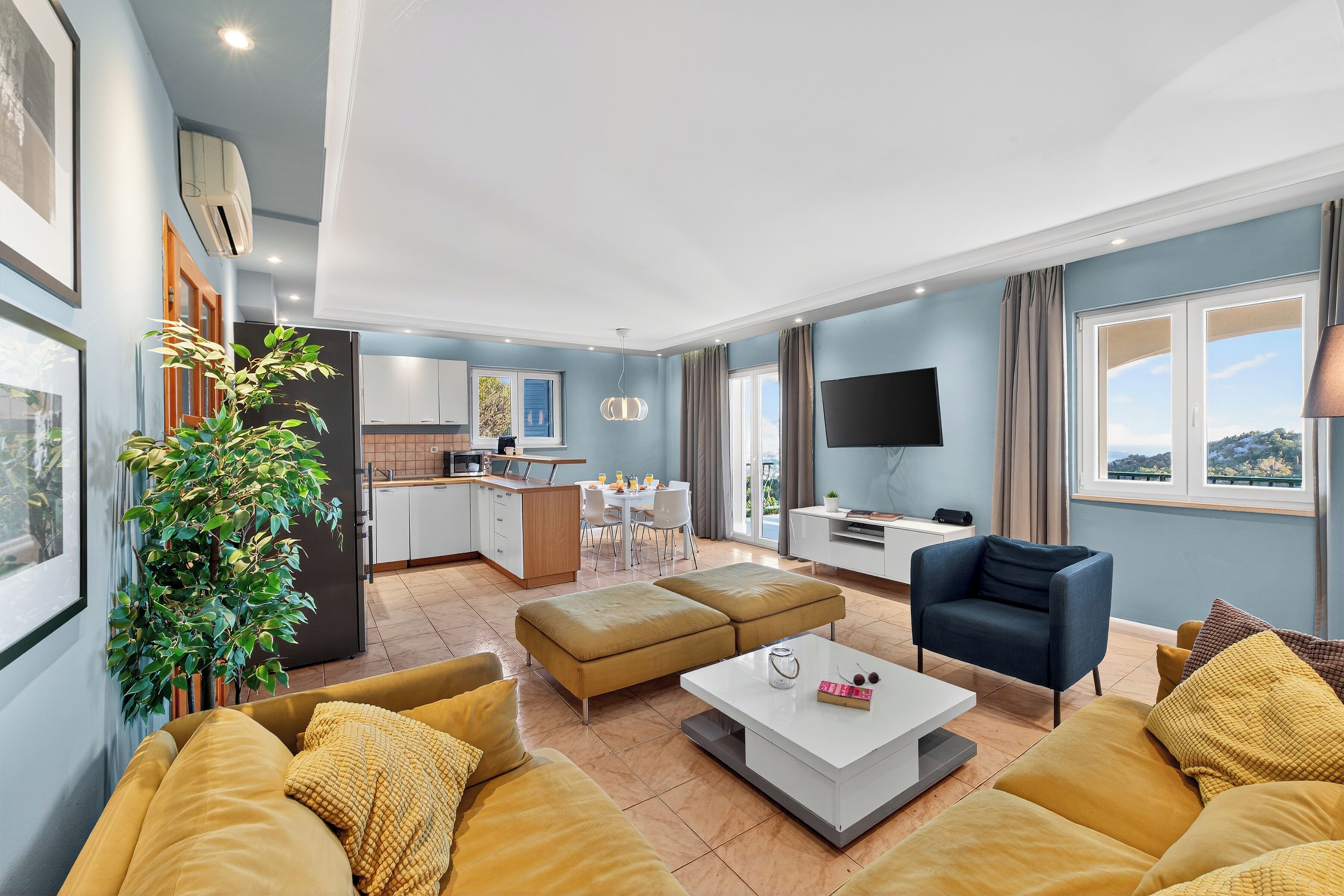 A modern living room with a sofa, ottoman, and a coffee table. There is a TV, a plant, and a view of the outside.