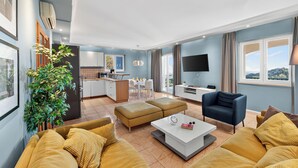 Een moderne woonkamer met een bank, poef en salontafel. Er is een televisie, een plant en uitzicht op buiten.