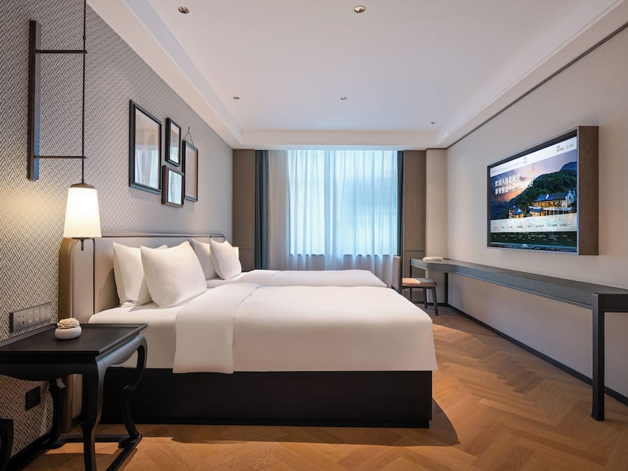 Quarto de hotel moderno com uma cama grande, mesa de cabeceira e televisão na parede.
