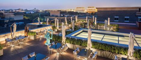 A rooftop terrace with tables and chairs, a pool, and a view of traditional buildings in the background.