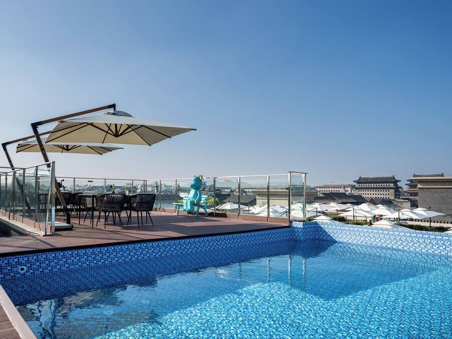 A rooftop pool with a clear view of the city skyline, a seating area with umbrellas, and a decorative frog statue.