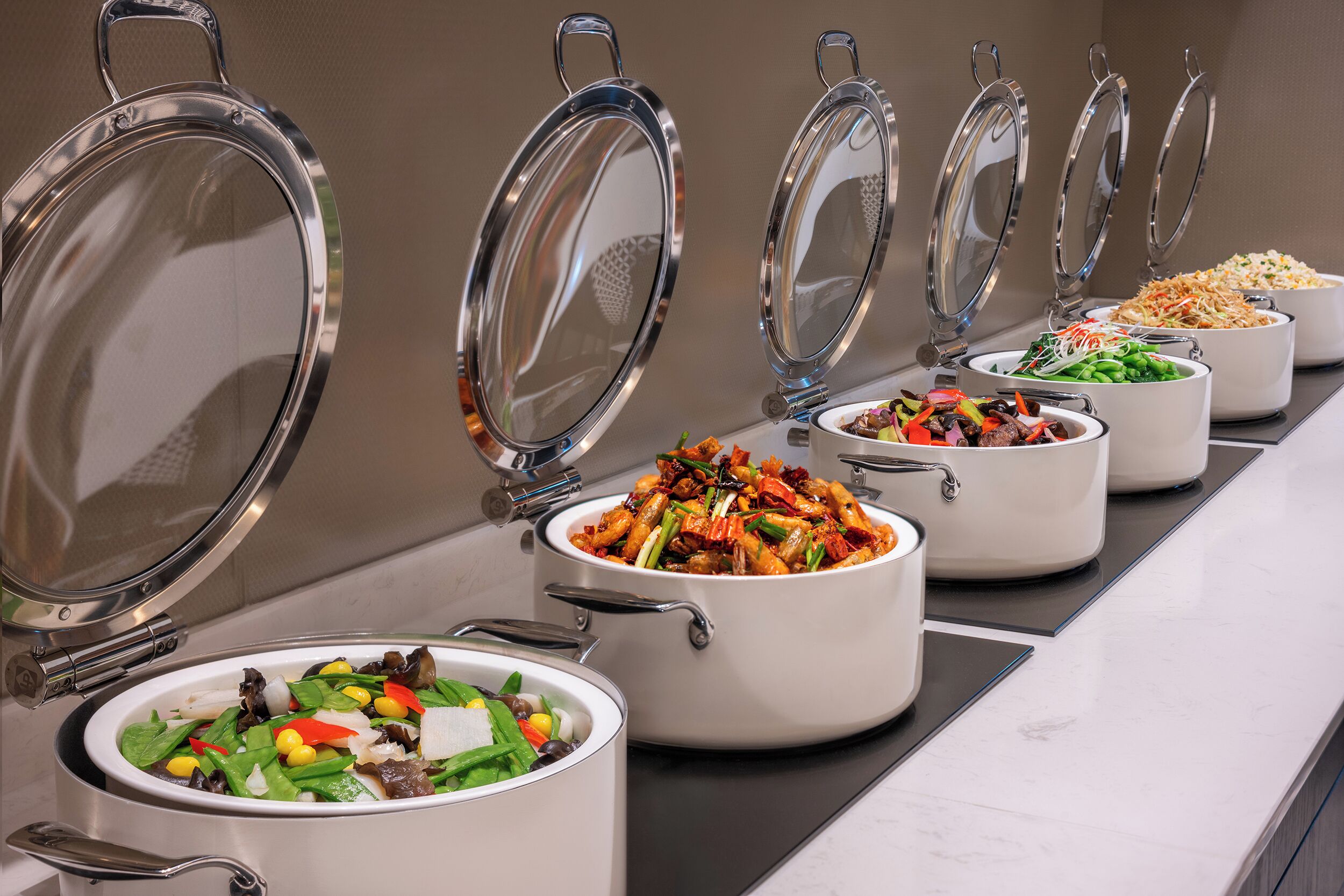 Una barra de buffet con diversos platos en ollas blancas, cada una con tapa metálica.