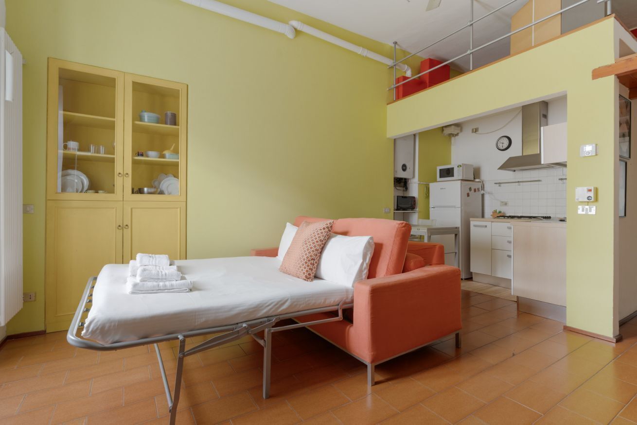 A room with a bed, a cabinet with dishes, a kitchenette, and a clock on the wall.