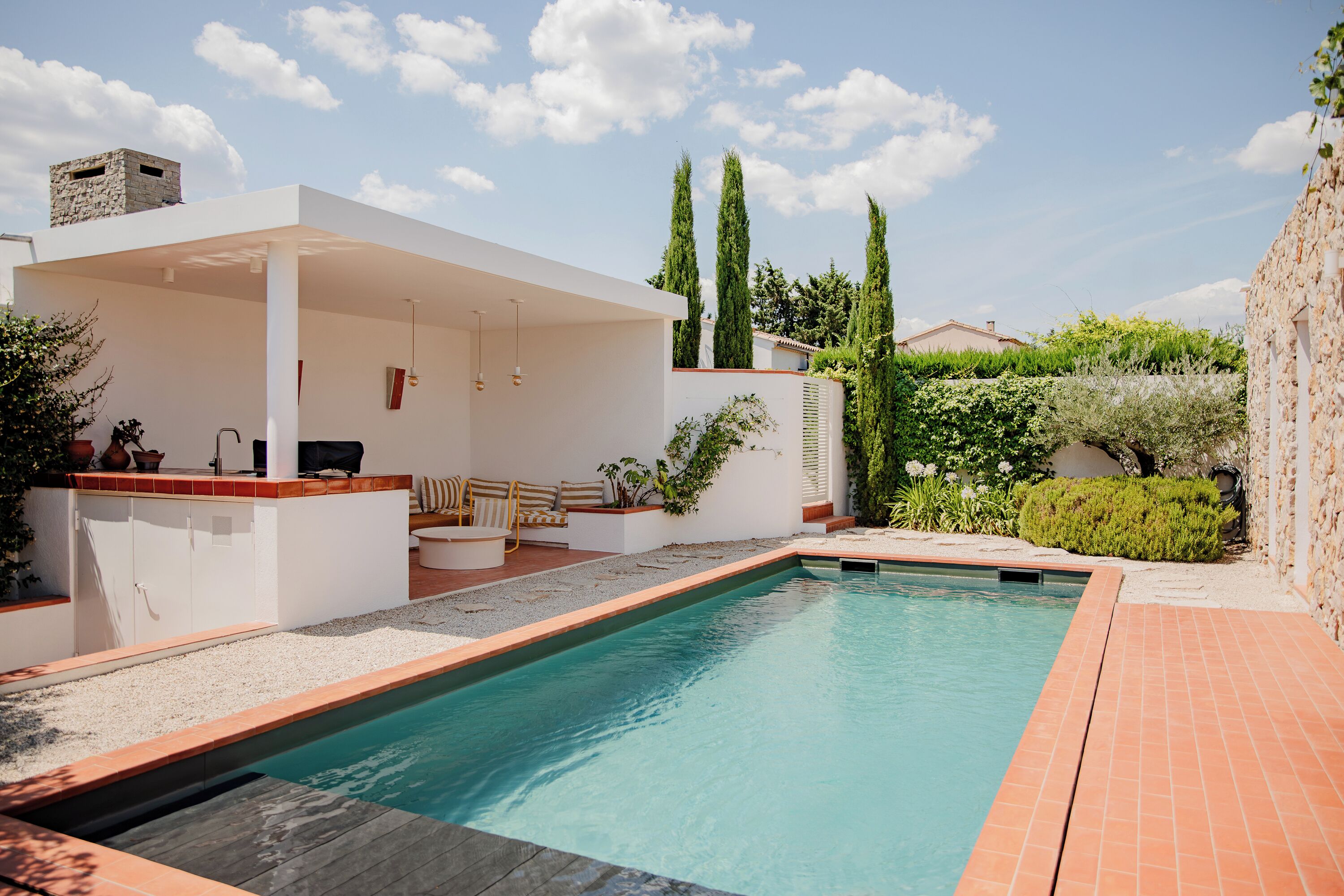 Un área de piscina moderna al aire libre con zona de estar cubierta, chimenea de piedra y abundante vegetación.