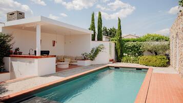 Un área de piscina moderna al aire libre con zona de estar cubierta, chimenea de piedra y abundante vegetación.