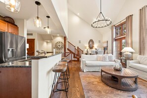 A spacious living area with a kitchen, wooden flooring, and a large window with curtains. - BLISS | Exclusive Tamarack Perks (Donnelly)