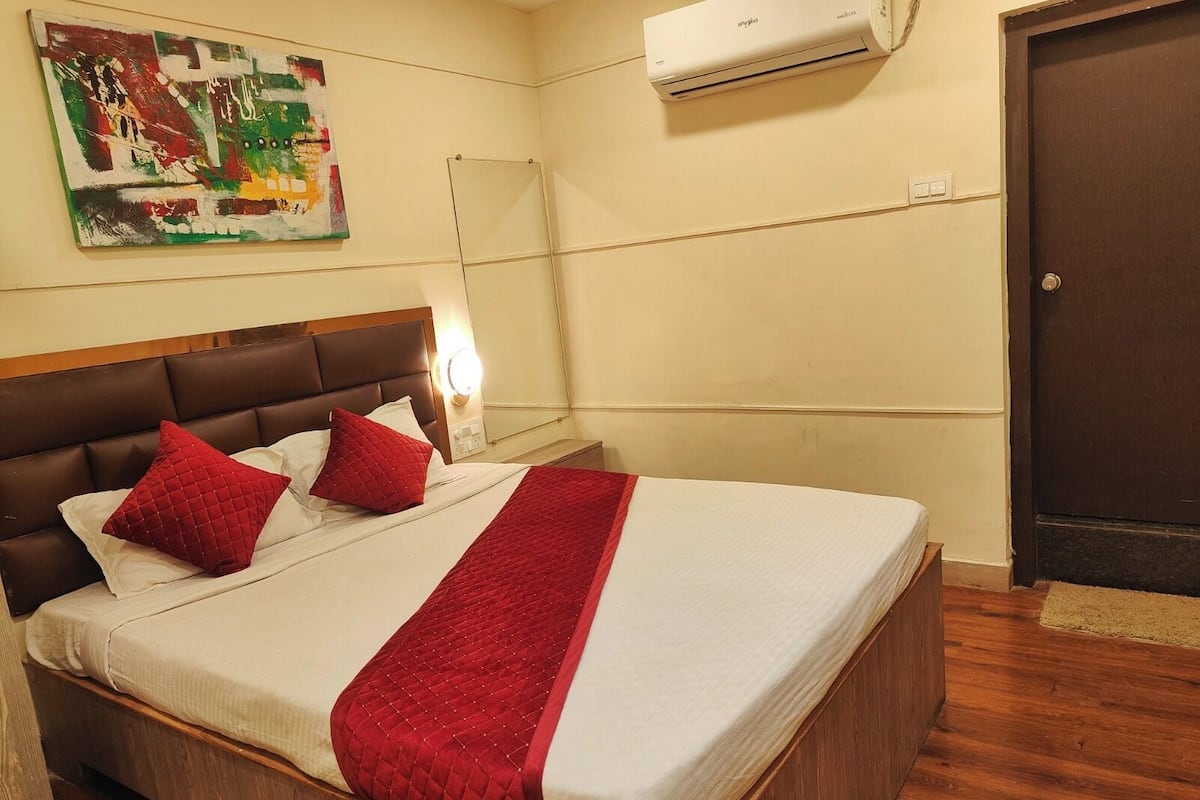Ein Hotelzimmer mit einem Bett, einer roten gesteppten Bettdecke, zwei roten Kissen, einem hölzernen Kopfteil, einer Wand-Klimaanlage und einem Bild an der Wand.