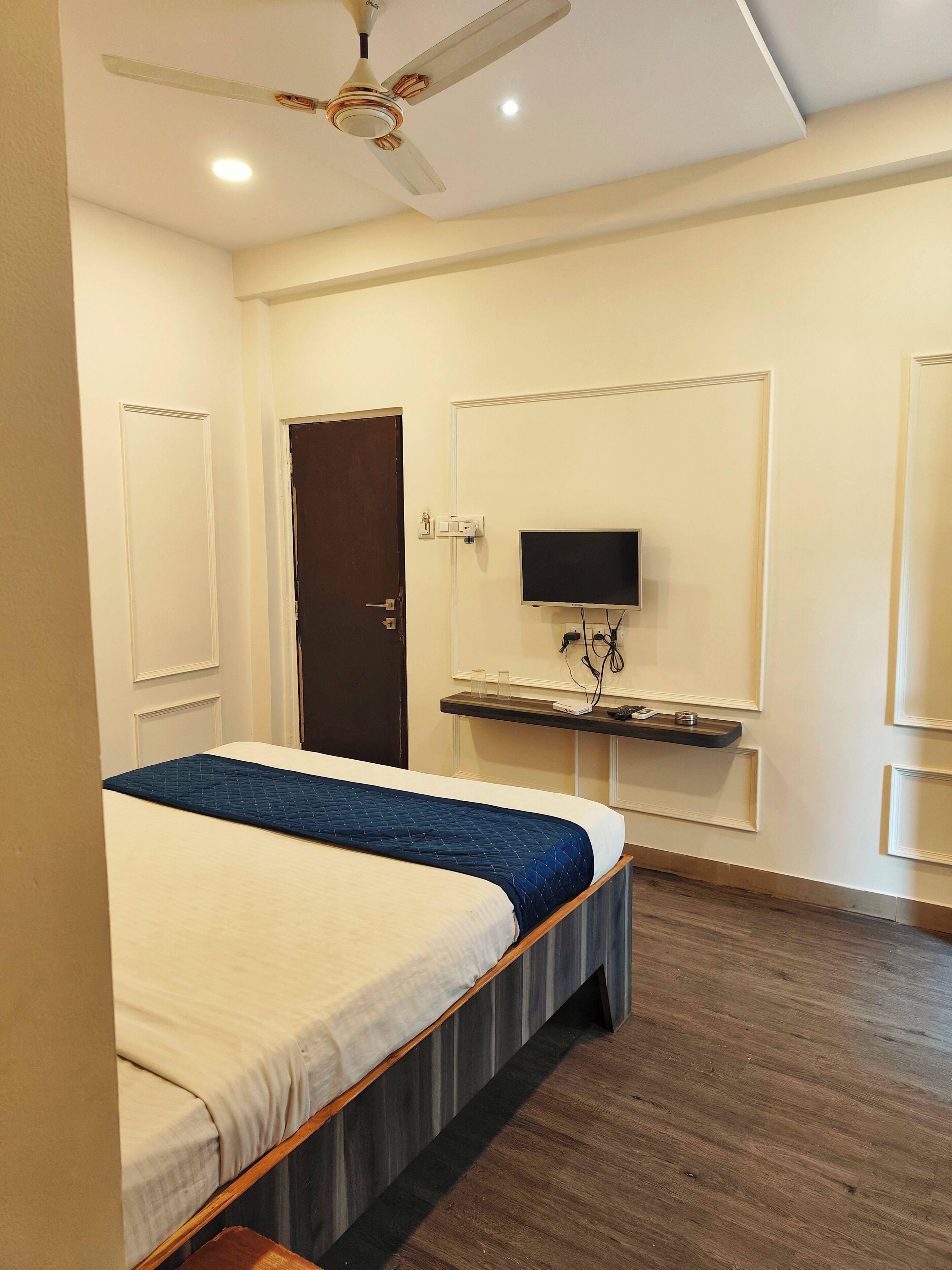 A hotel room with a bed, a desk with a flat-screen TV, a ceiling fan, and a door.