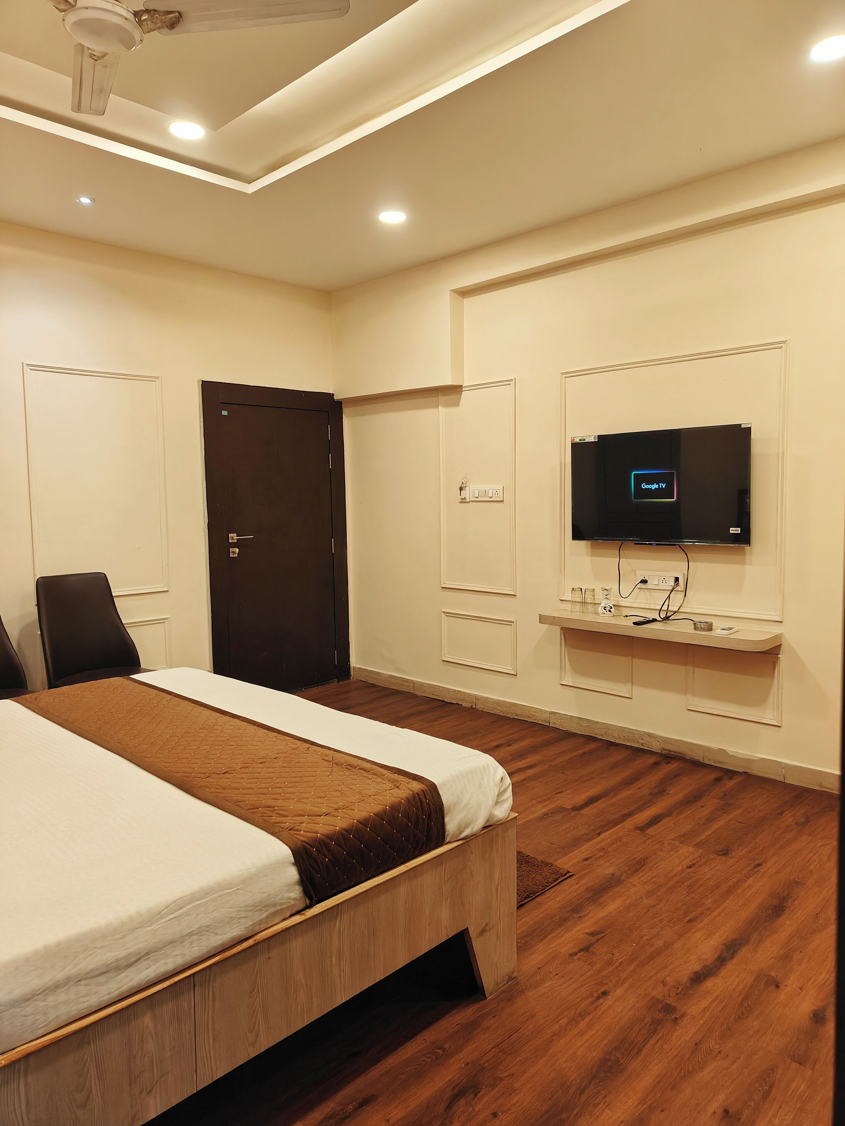 Una habitación de hotel con una cama, un televisor montado en la pared, piso de madera y ventilador de techo.