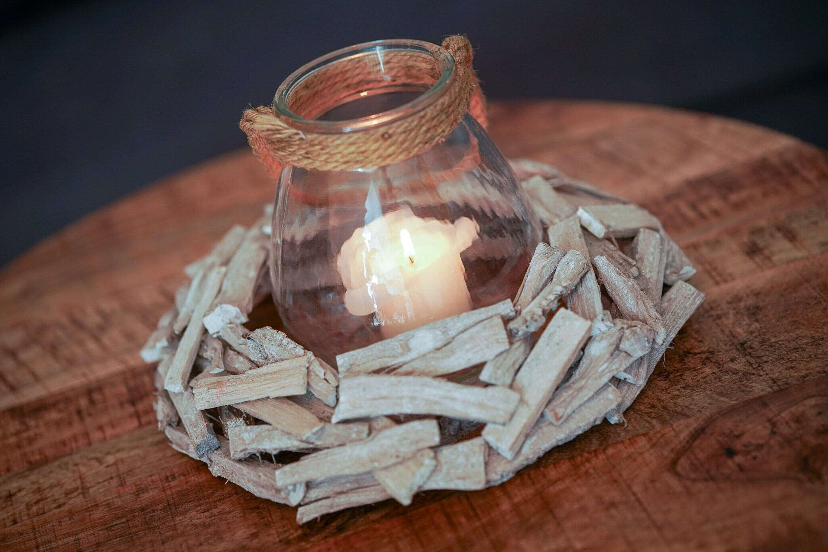 A glass candle holder with a rope handle, surrounded by wood shavings, on a wooden surface.