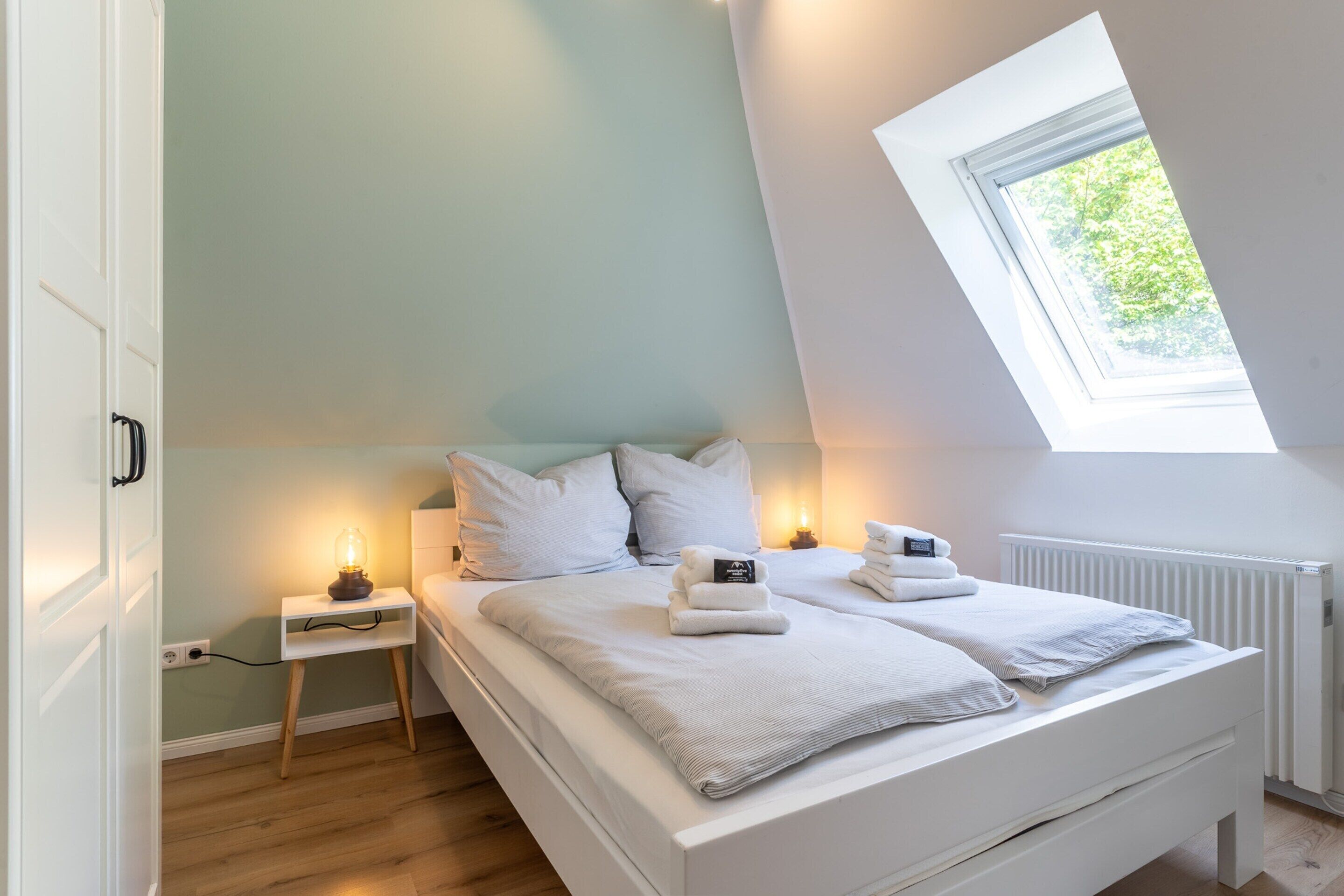Quarto com teto inclinado, claraboia, cama com roupa de cama branca, mesa de cabeceira com abajur e radiador.