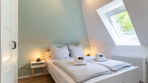 A bedroom with a sloped ceiling, a skylight, a bed with white bedding, a bedside table with a lamp, and a radiator. - Ferienwohnung mit Balkon in St.peter-ording (St. Peter-Ording)