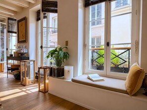 Ein Zimmer mit großem Fenster, einer Bank mit Kissen, einem Blumentopf, einer Kerze und einem kleinen Tisch mit Lampe.