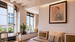 A cozy living room with a large window, a sofa with cushions, a framed artwork on the wall, and a wooden floor. - L'appartement Charlot C (Paris)