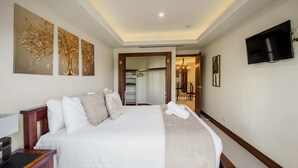 Een keurig opgemaakt bed met witte lakens en een beige plaid, een houten nachtkastje met een lamp, en een flatscreen televisie aan de muur.