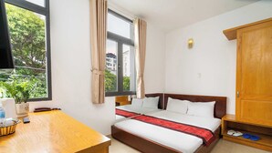 Een hotelkamer met een bed, een bureau, een raam met gordijnen, een houten kledingkast en een klein plankje met slippers.