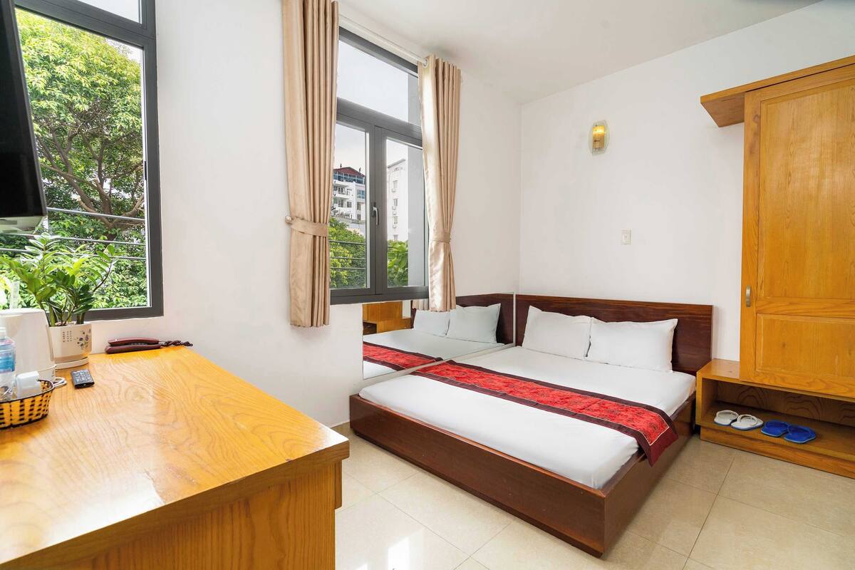 Een hotelkamer met een bed, een bureau, een raam met gordijnen, een houten kledingkast en een klein plankje met slippers.