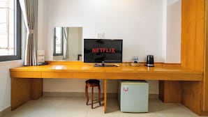 Een compacte kamer met een houten bureau, een televisie waarop 'NETFLIX' wordt weergegeven, een kleine koelkast en een raam met gordijnen.