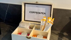 En trææske med rum til kaffeposer og teposer, et indrammet skilt med teksten 'COFFEE&TEA' og 'CAFÉ Y TÉ', samt en lille rød genstand.