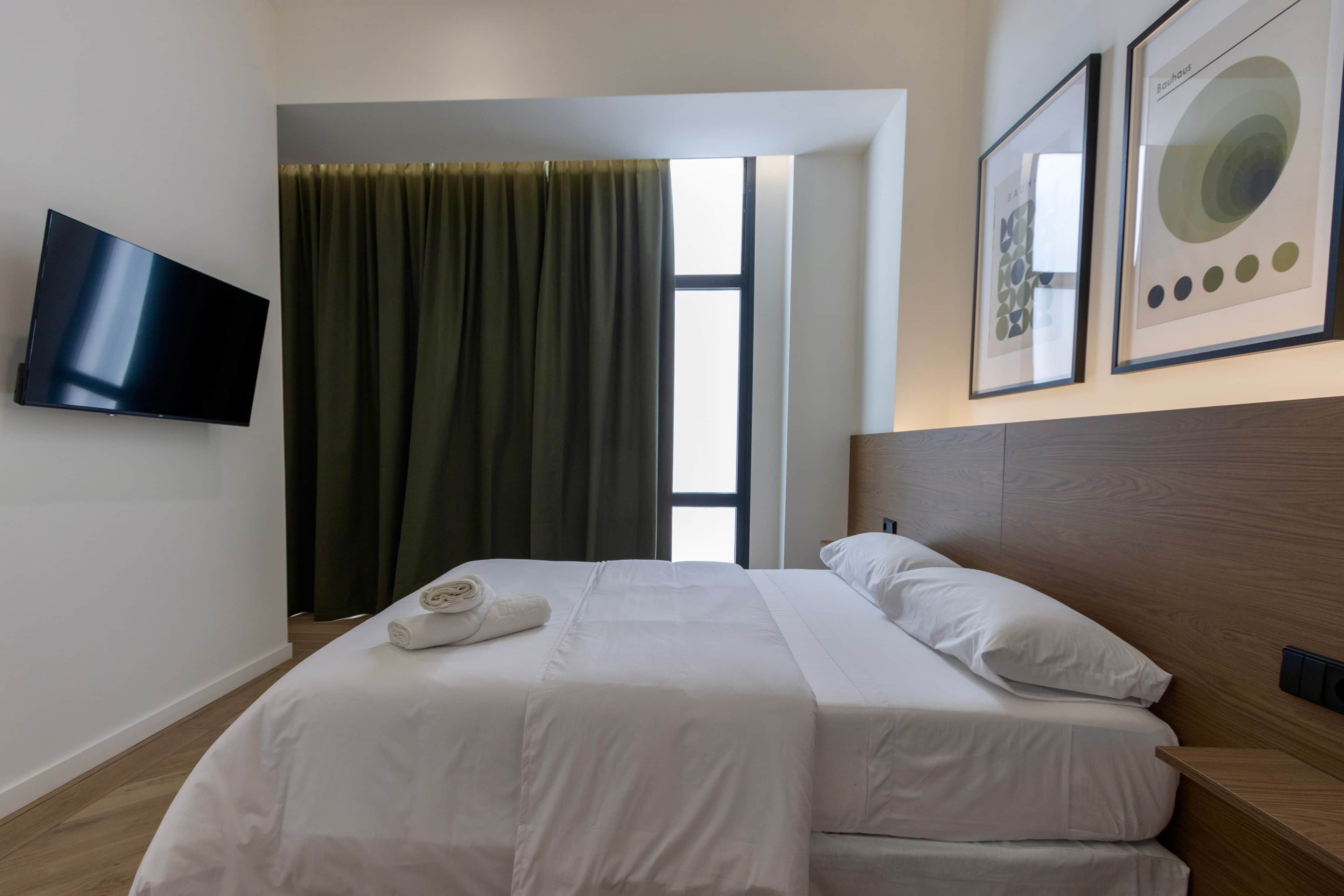 Une chambre d’hôtel avec un lit, un téléviseur fixé au mur, une fenêtre avec des rideaux et deux œuvres d’art encadrées accrochées au mur.