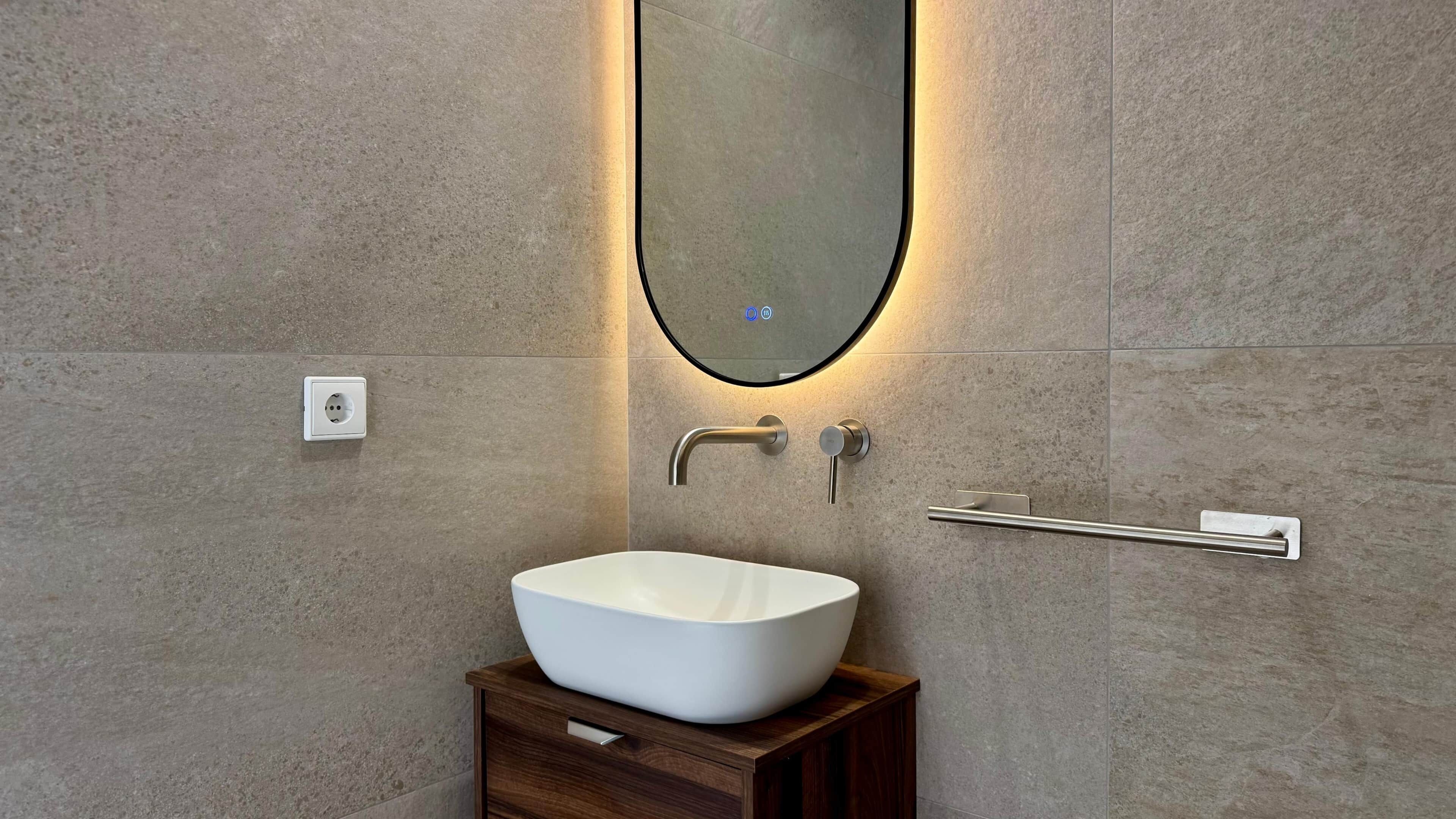 Un baño con un lavamanos blanco tipo lavabo, un mueble de tocador de madera, un espejo montado en la pared y un perchero para toallas.