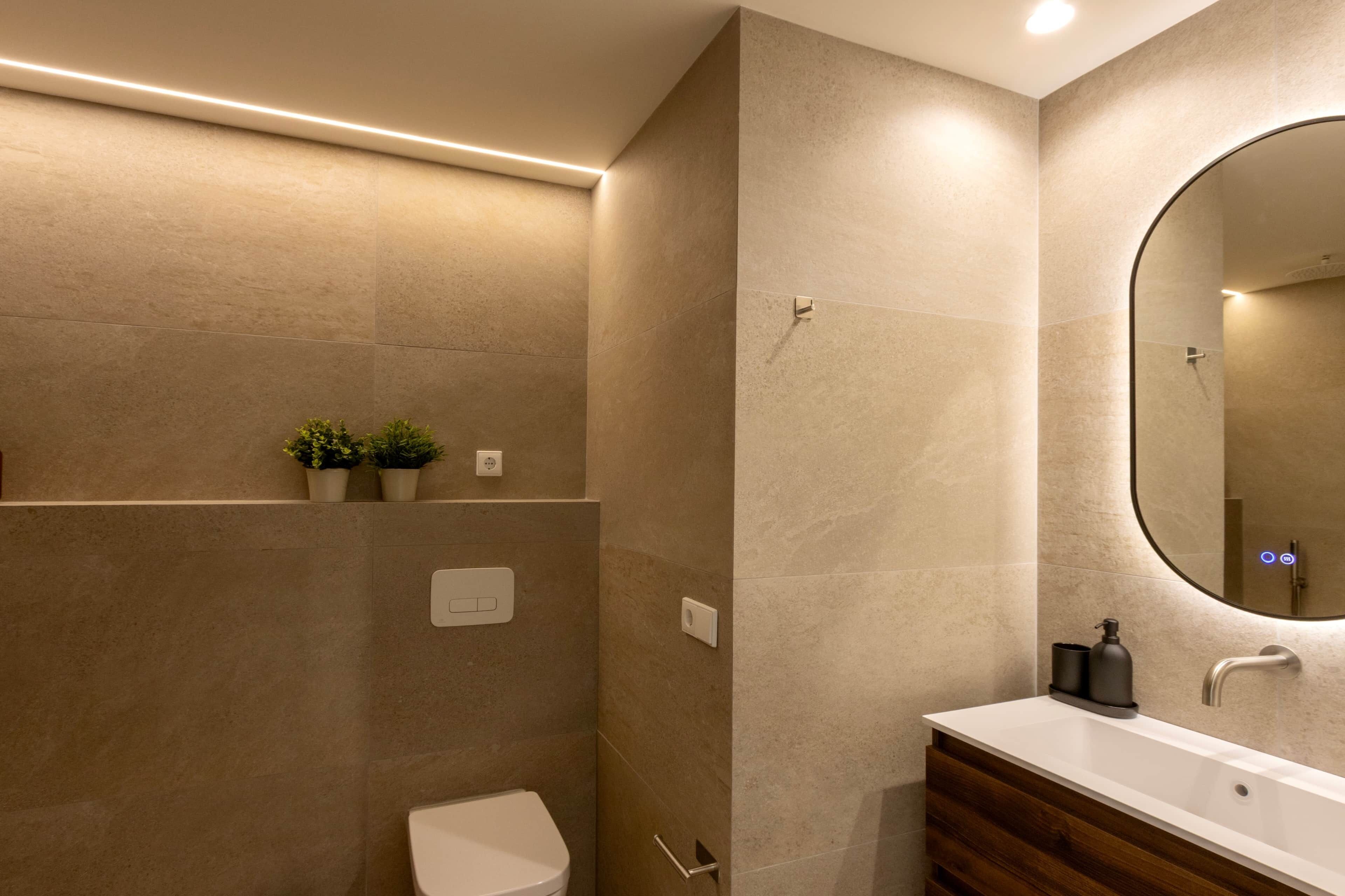 Un baño moderno con inodoro blanco, tocador de madera, espejo grande y estante de pared con plantas.