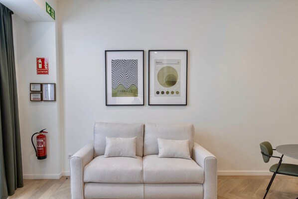 A modern room with a light-colored sofa, two framed artworks, a fire extinguisher, and a small table with a chair. - One Tower Apartment - 07 (Valencia)