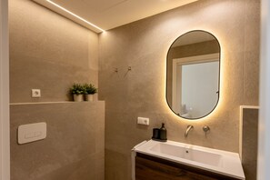 Un baño moderno con un lavabo blanco, un espejo grande con iluminación debajo y una pequeña planta en maceta en la pared.