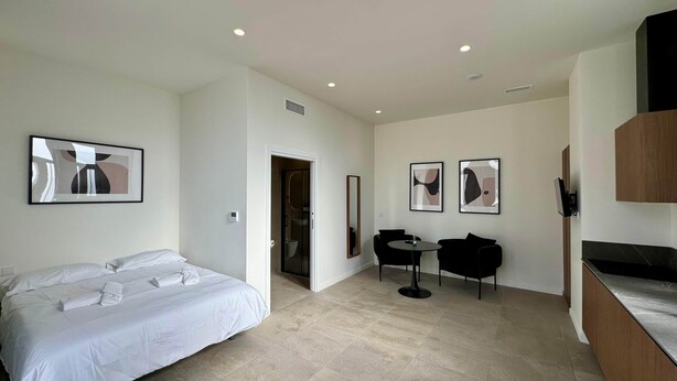 Una habitación de hotel moderna con cama, una pequeña mesa de comedor y dos sillas. Hay una puerta que da al baño, y la habitación tiene un diseño limpio y minimalista.