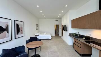 Una habitación de hotel moderna con cama, una pequeña zona de estar, una kitchenette y un baño visible a través de una puerta abierta.