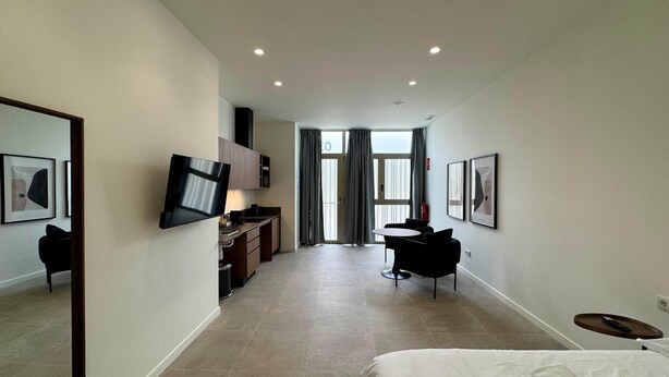 A modern hotel room with a bed, a TV mounted on the wall, a desk with a chair, and a dining area with a table and chairs.