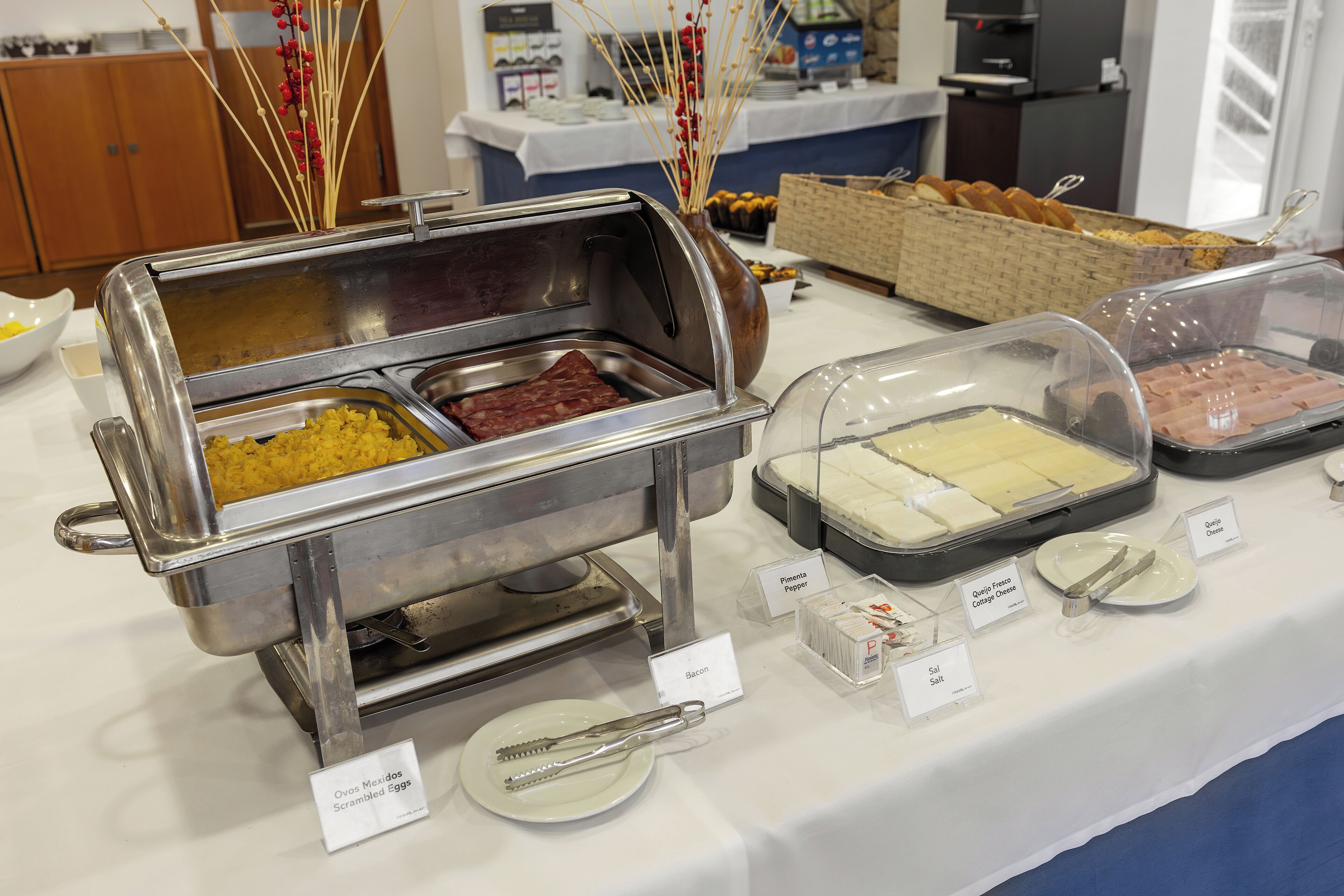 Un buffet proposant une variété de plats, chacun étant clairement identifié, notamment des œufs brouillés, du bacon, du fromage frais et du jambon.
