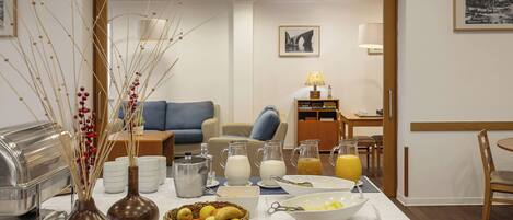 Montaje de un bufé de desayuno con diferentes alimentos, bebidas y un área de comedor con sofá y mesa.