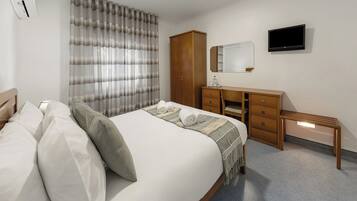 A hotel room with a bed, a wooden wardrobe, a desk with drawers, a mirror, and a flat-screen TV mounted on the wall.